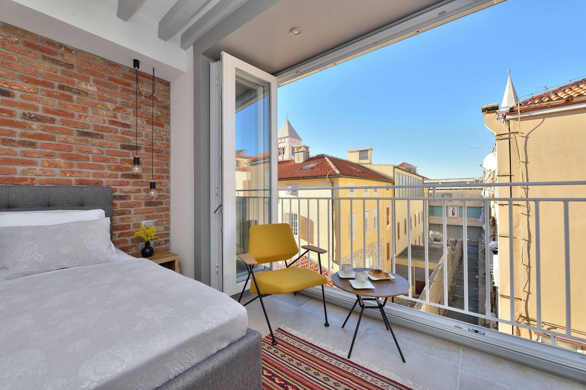 A modern bedroom with a brick wall, a bed, a yellow chair, and a balcony with a city view.