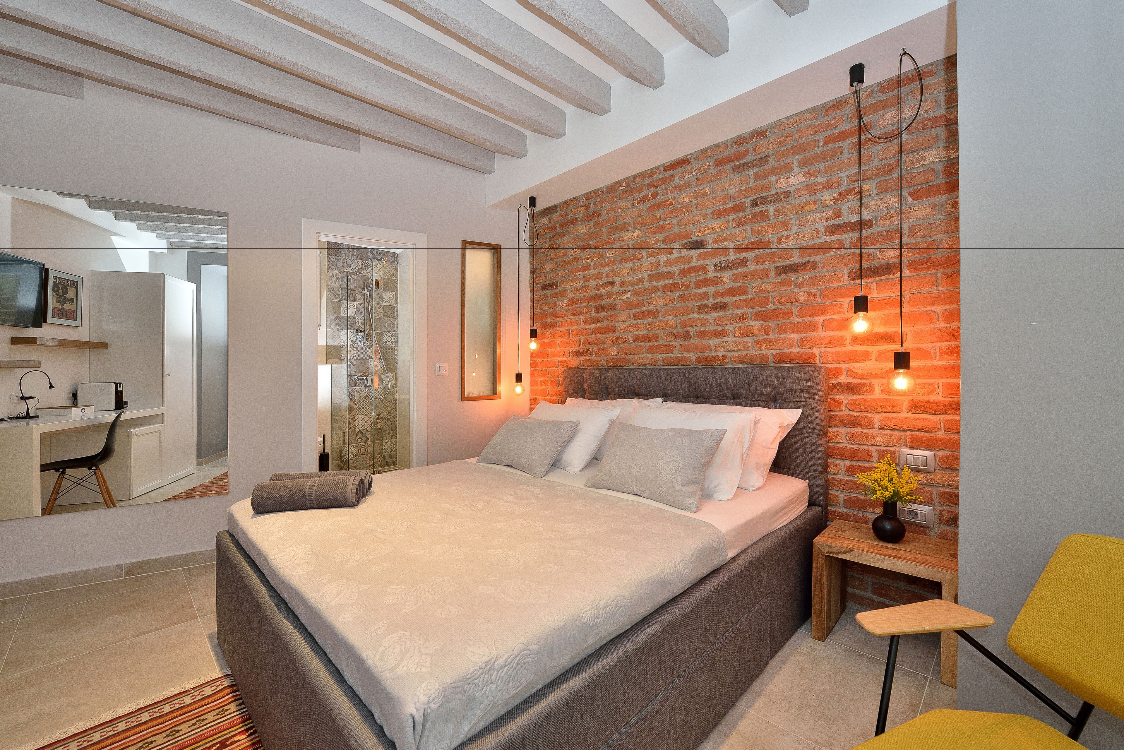 Une chambre moderne avec un mur en briques, un lit doté d’une tête de lit grise, un petit bureau et une chaise jaune.