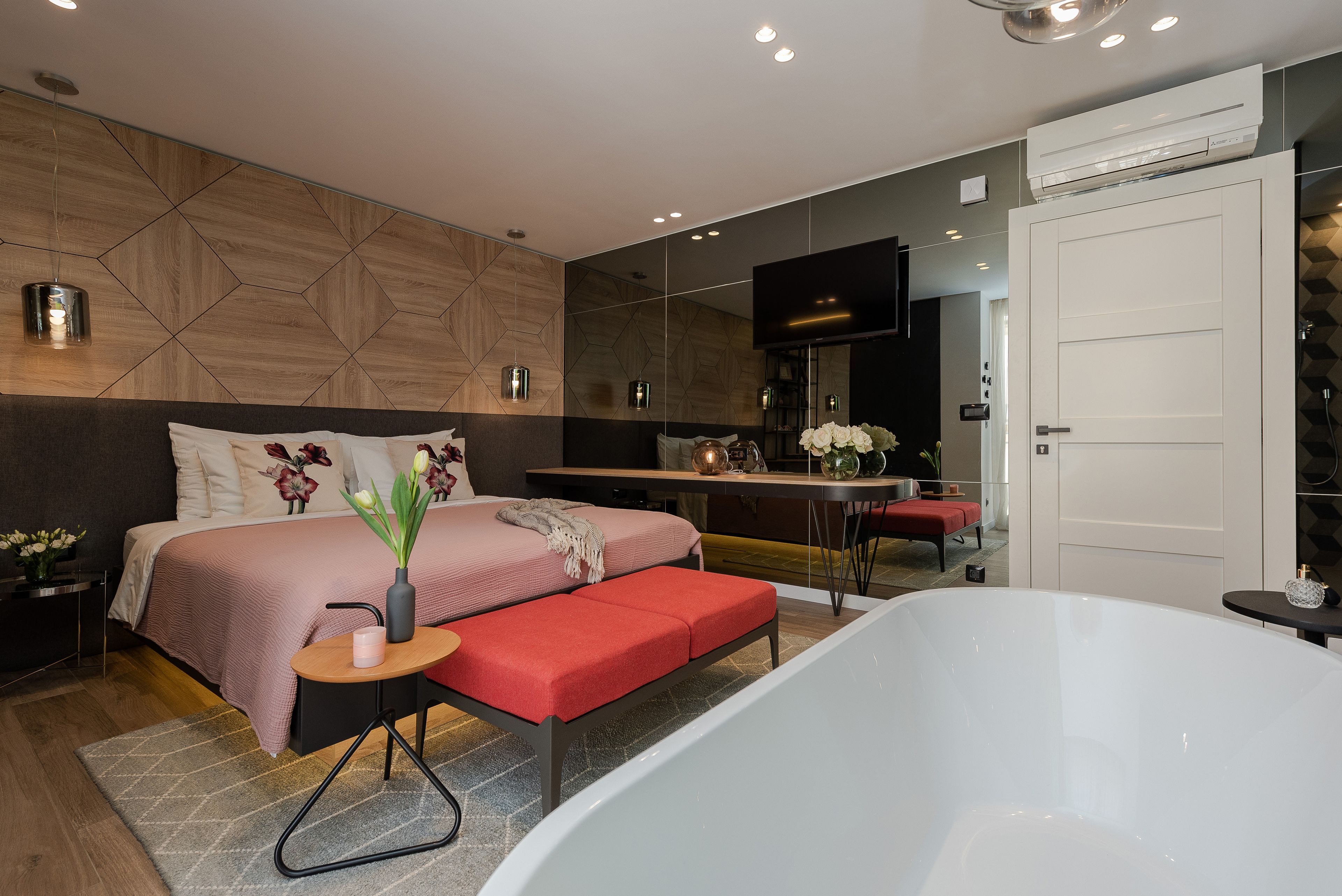 Un dormitorio moderno con una cama grande, un banco rojo, una bañera blanca y una pequeña mesa con un florero.
