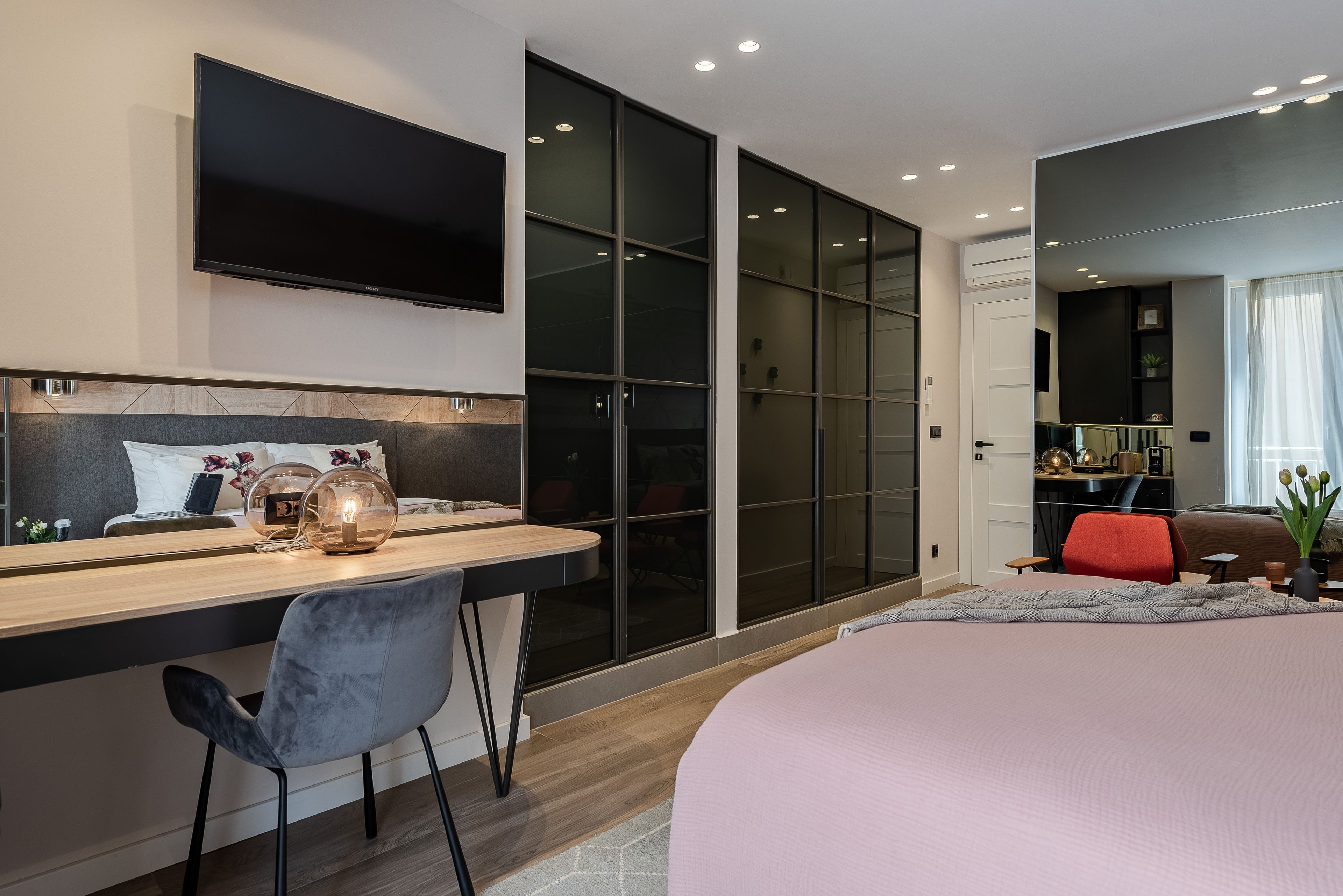 A modern bedroom with a large bed, a desk with a chair, a flat-screen TV, and a sliding glass door leading to a balcony.