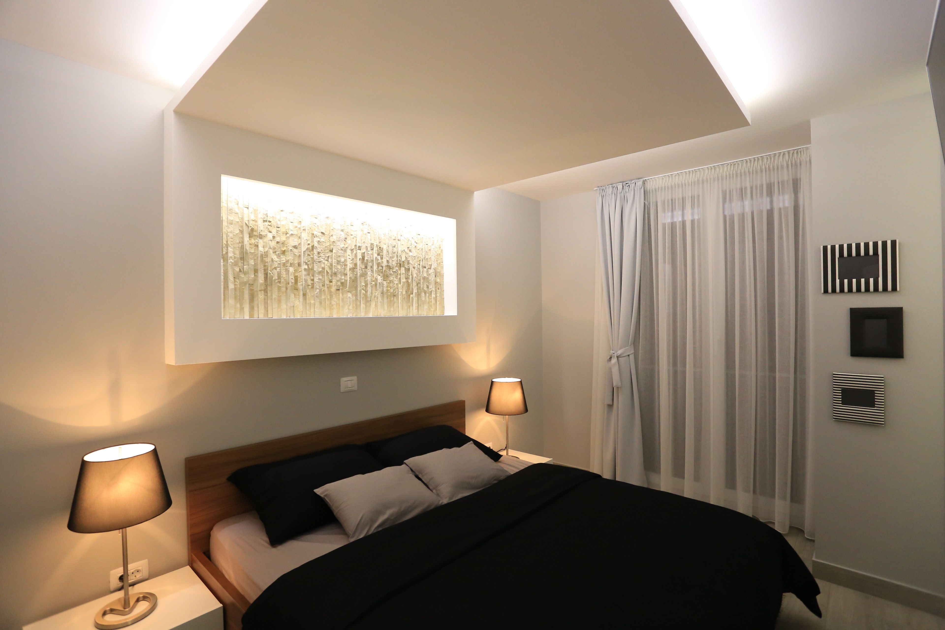 Une chambre moderne avec un lit, deux lampes de chevet, une fenêtre avec des rideaux légers et une œuvre d’art accrochée au mur.