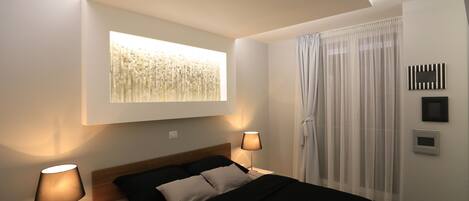 A modern bedroom with a bed, two bedside lamps, a window with sheer curtains, and a wall-mounted artwork.