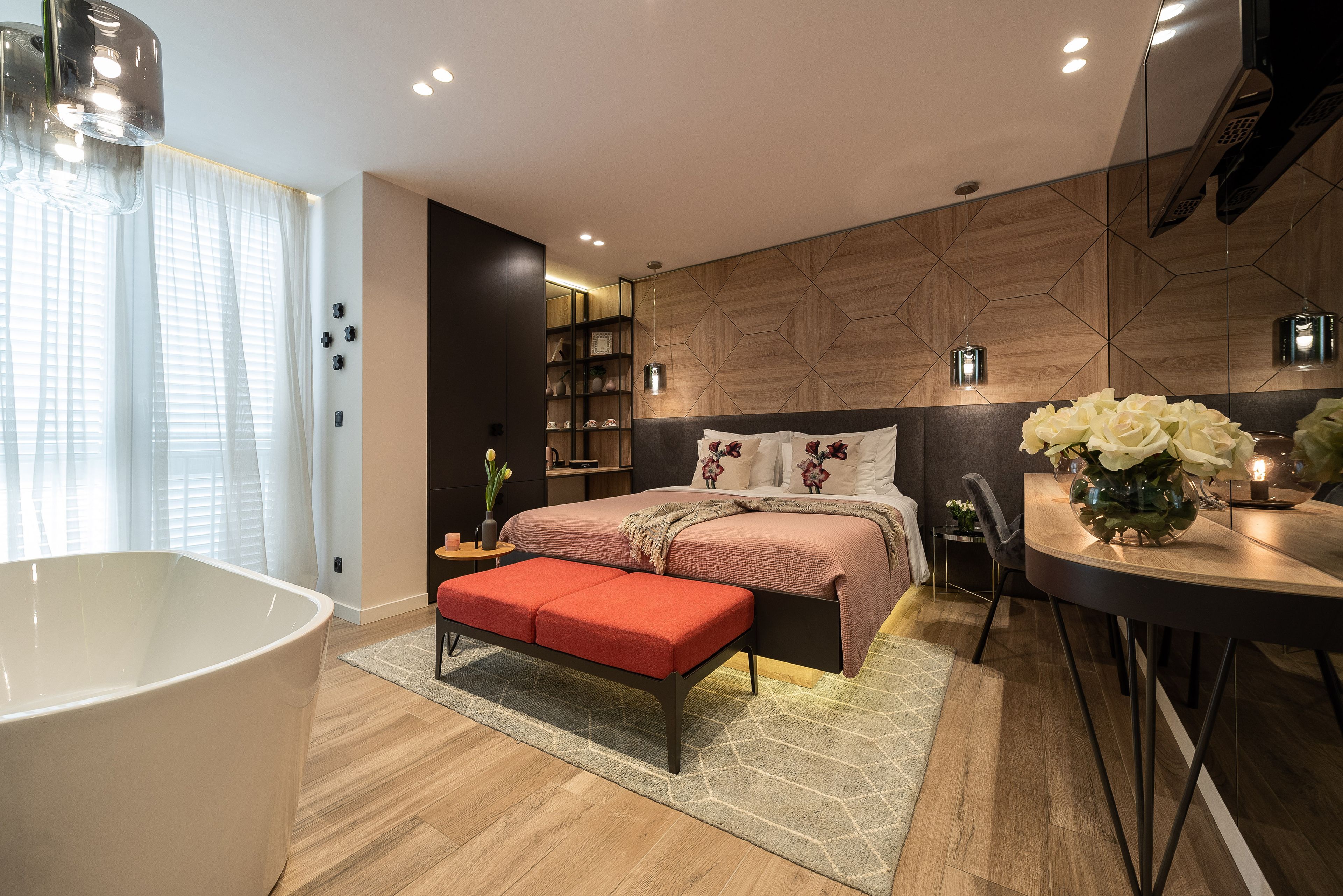 Un dormitorio moderno con una cama grande, una mesita de noche, un banco rojo, una bañera blanca y un panel de pared de madera.