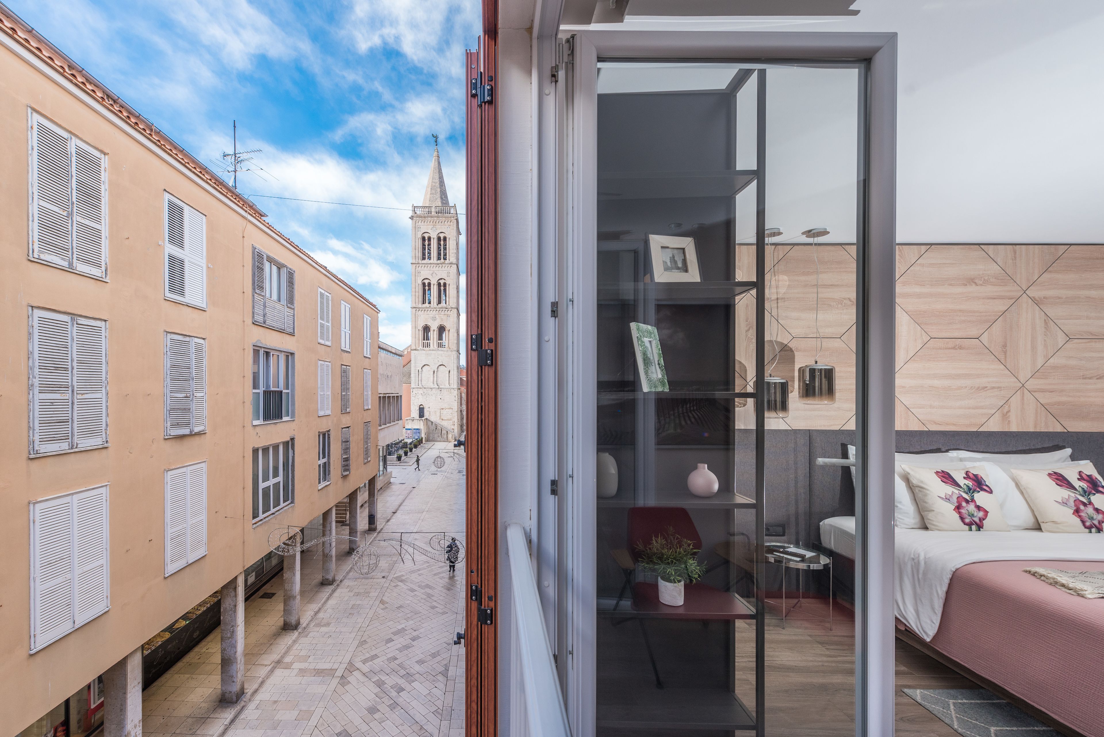 En moderne lejlighed med en glasdør, der fører til et soveværelse med seng og et lille bord. Der er en balkon med udsigt til et historisk tårn.