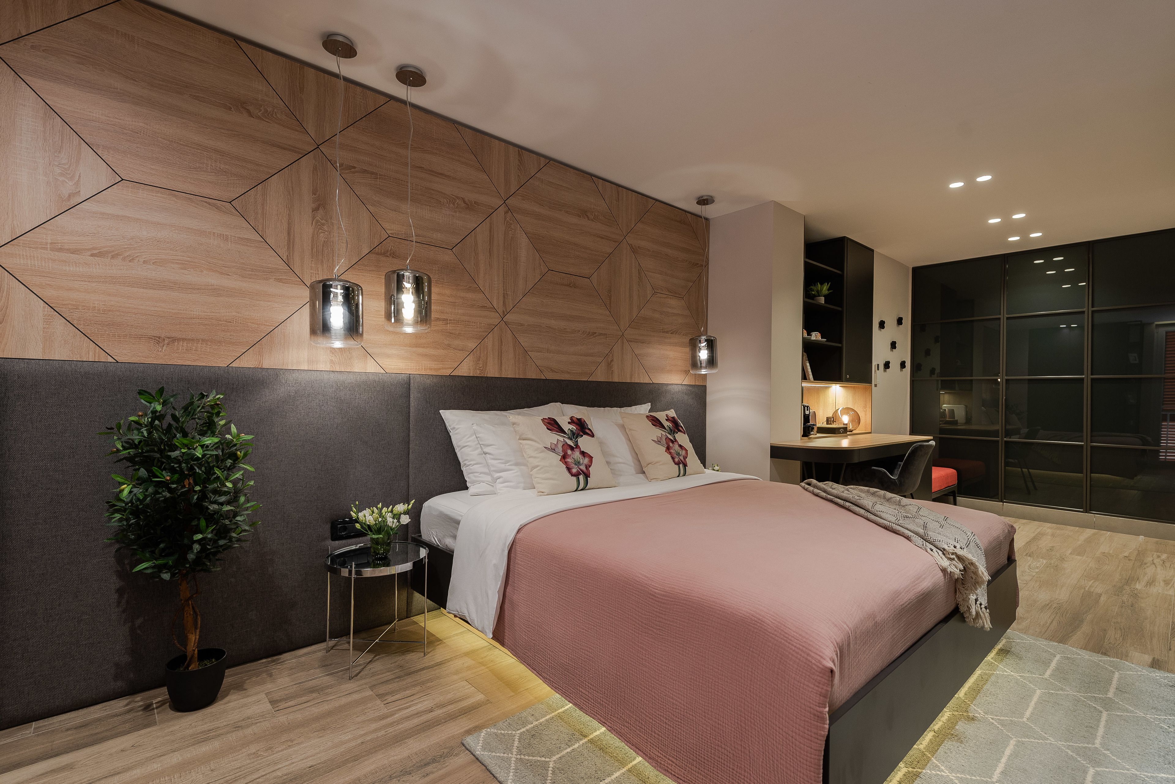 Un dormitorio moderno con una cabecera de patrón geométrico, una cama con colcha rosa, una mesita de noche con lámpara y una planta en maceta.