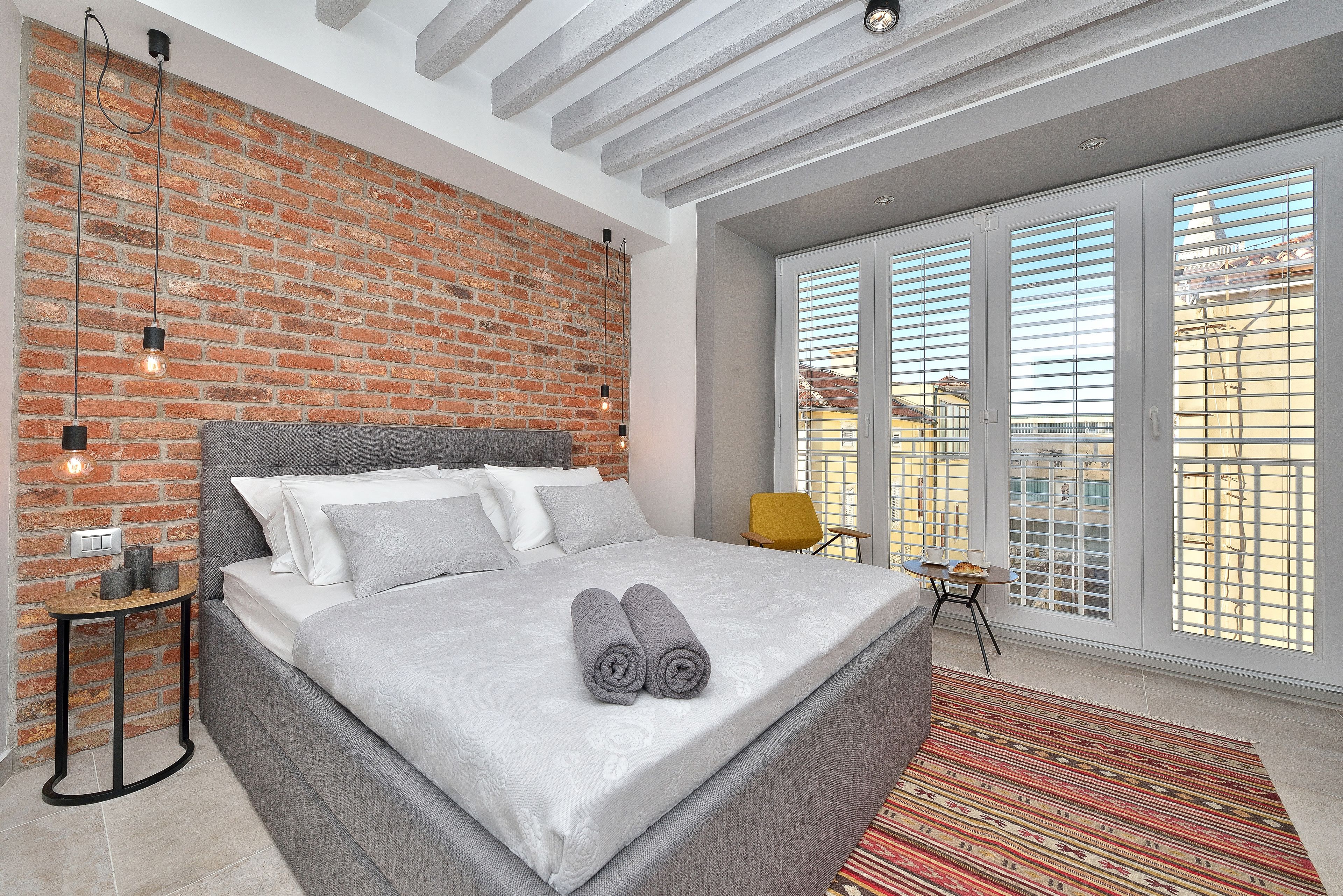 A modern bedroom with a brick wall, a large bed with grey bedding, a small table, and a view through large windows.