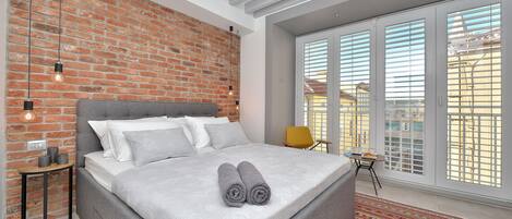A modern bedroom with a brick wall, a large bed with grey bedding, a small table, and a view through large windows.