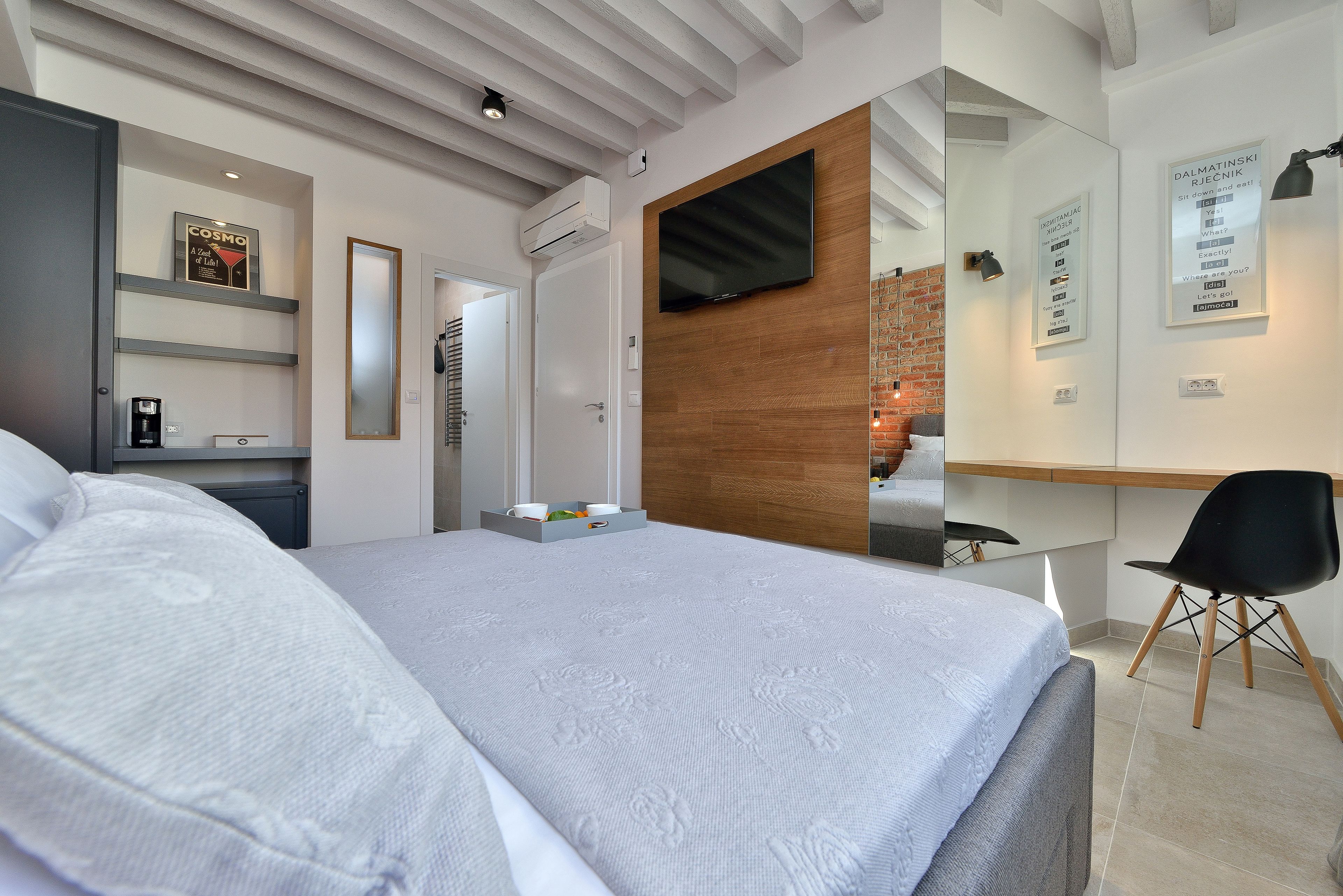 A modern hotel room with a large bed, a desk with a chair, a TV mounted on a wooden wall, and a mirror.