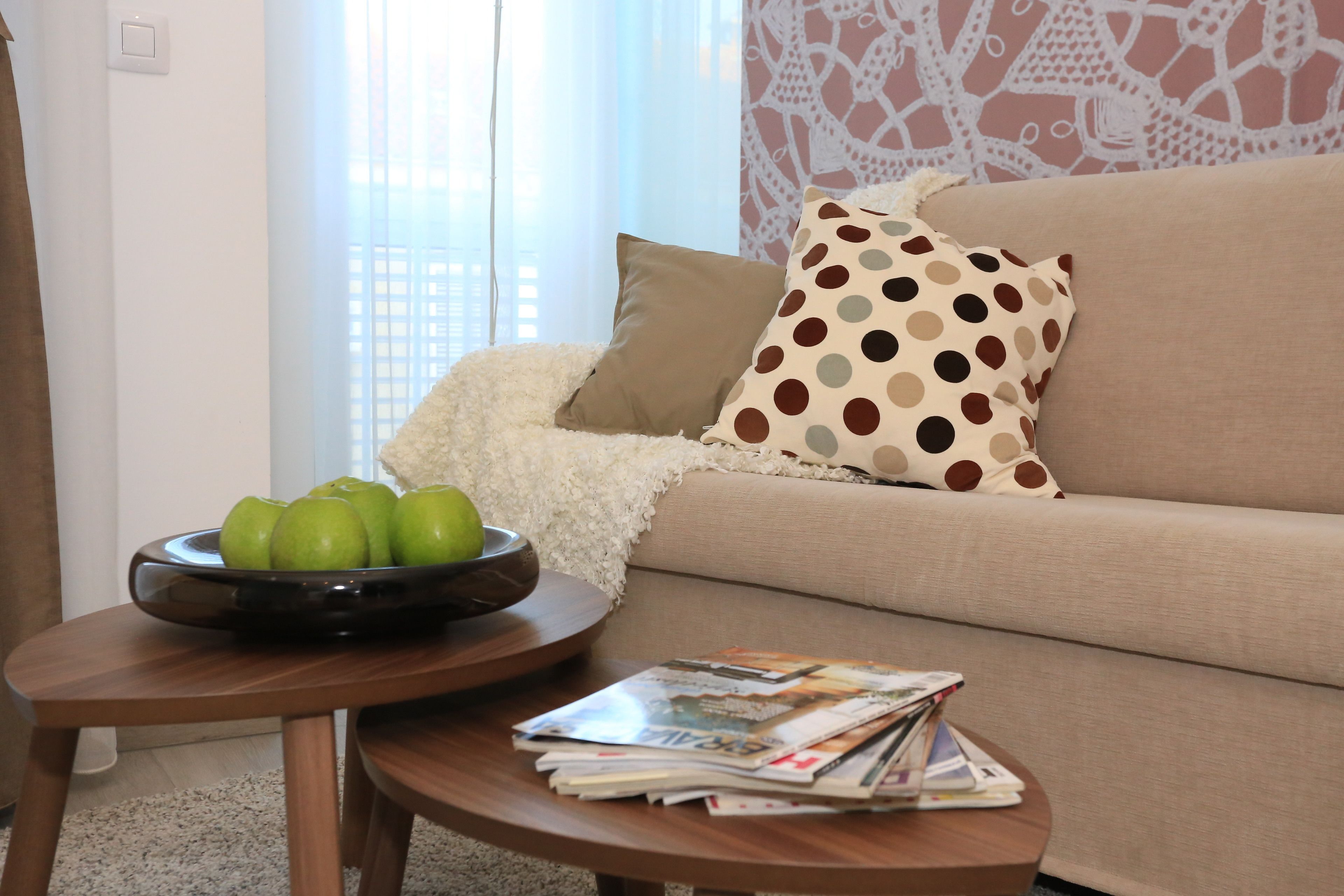 A living room with a sofa, a round wooden table with a bowl of green apples, and a stack of magazines.