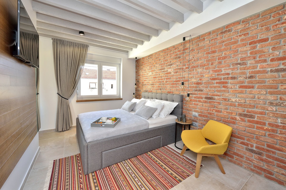 A modern bedroom with a brick wall, a grey bed, a yellow chair, and a window with curtains.
