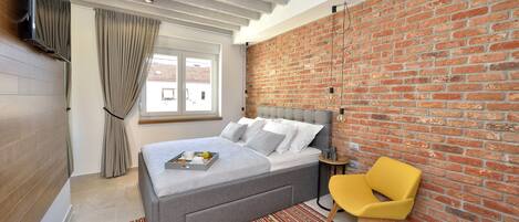 Une chambre moderne avec un mur en briques, un lit gris, une chaise jaune et une fenêtre avec des rideaux.