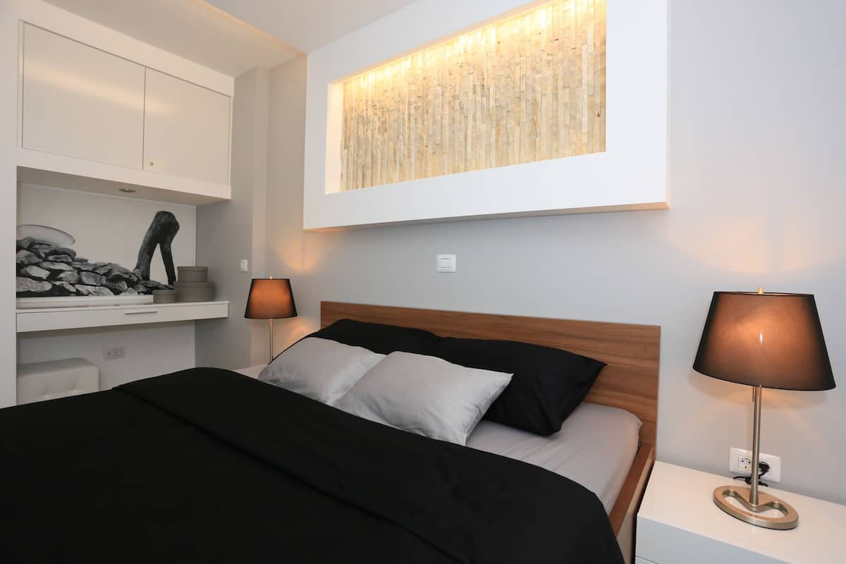 A neatly arranged bedroom with a bed, bedside lamps, a wall-mounted artwork, and a built-in shelf with decorative items.