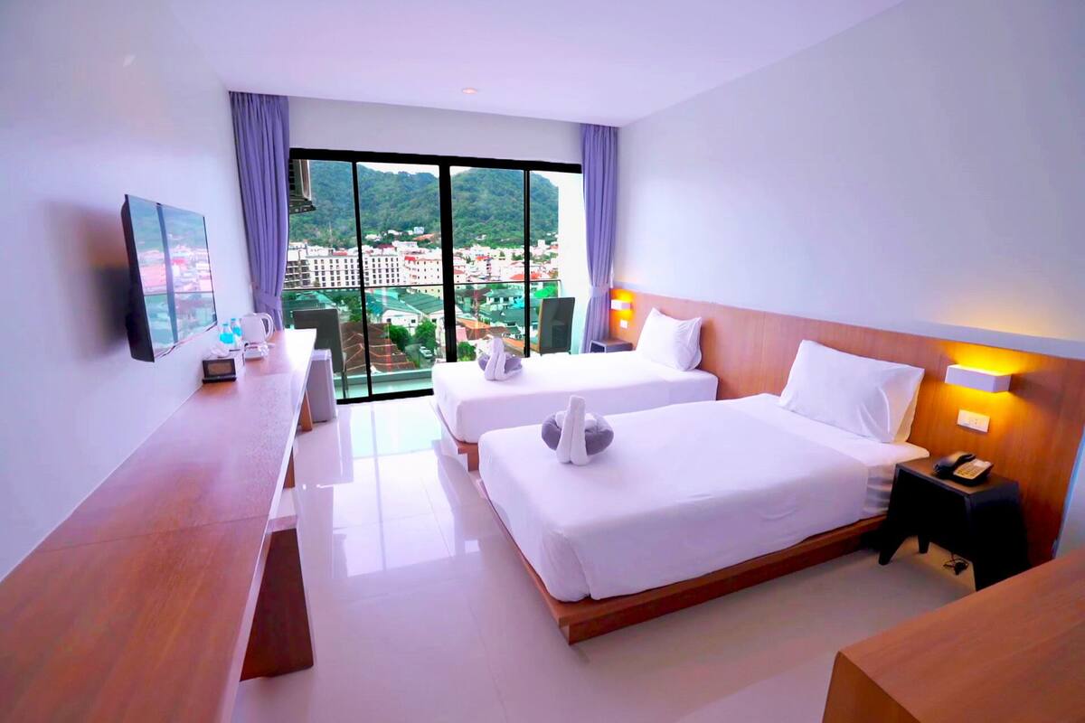A hotel room with two beds, a desk, a chair, a TV, and a large window with a city view.