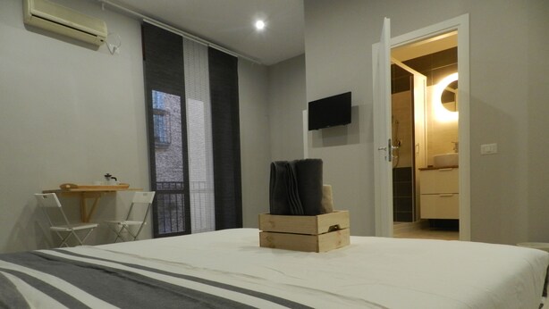A bedroom with a bed, a desk, and a chair. There is a window with blinds, a flat-screen TV on the wall, and a wooden box with towels on the bed. - Mola Rooms (Naples)
