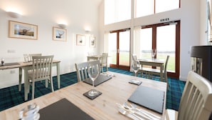A dining area with a wooden table, chairs, and a view of the outdoors through large windows. - The Steading (Dornoch)