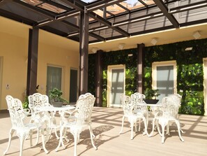 An outdoor patio with white ornate chairs and tables, a green wall with plants, and a metal roof structure. - Aeterno Eco Guest House (Rome)