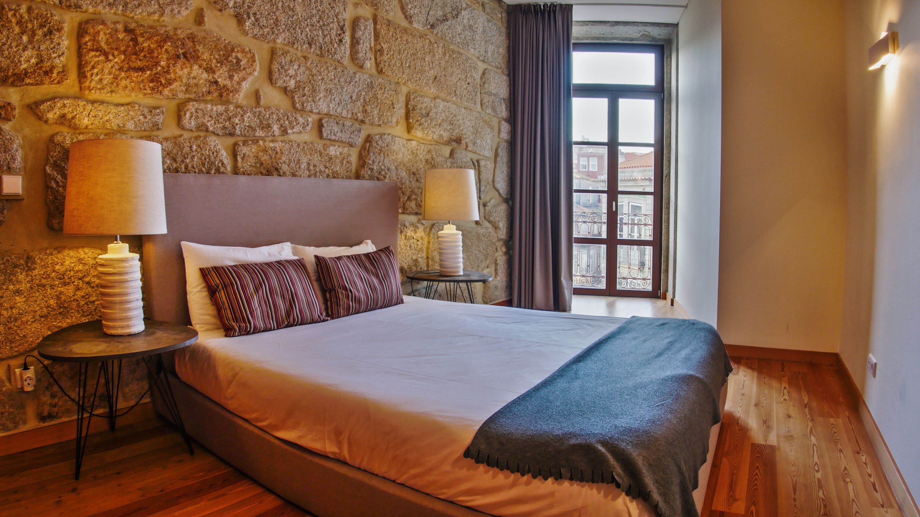 A hotel room with a stone wall, a large bed with white linens, two bedside lamps, and a view of buildings through a window.