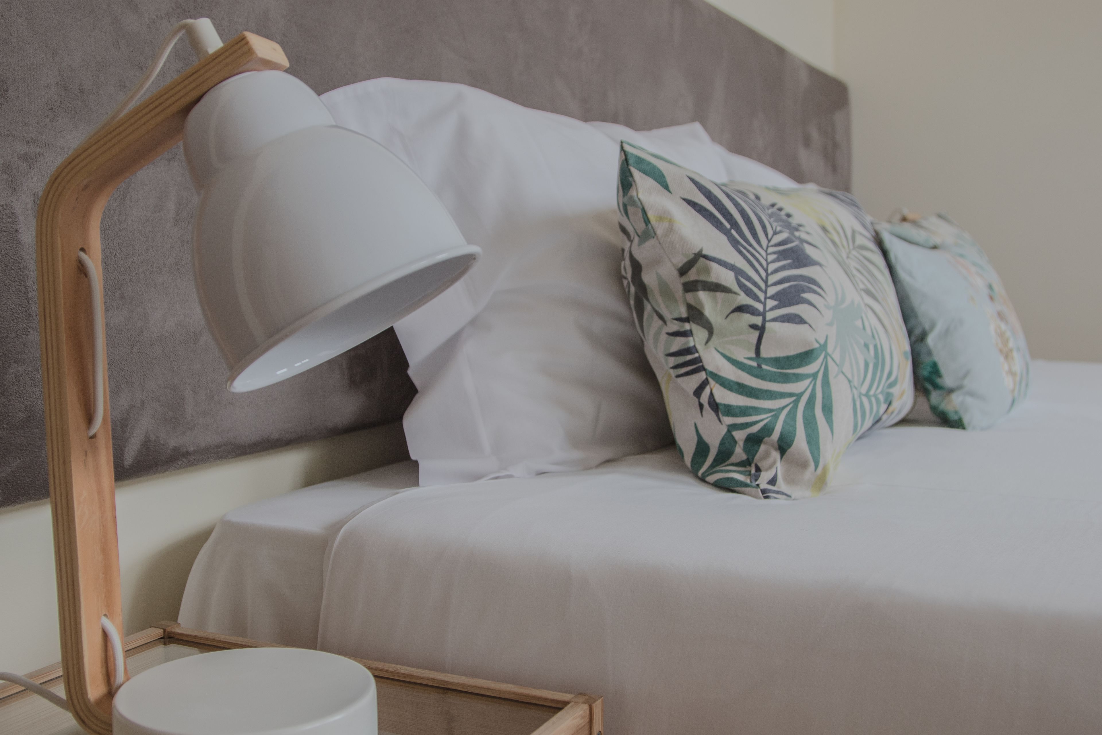 A neatly made bed with white bedding and a leaf-patterned pillow, a wooden bedside table with a white lamp, and a grey headboard.