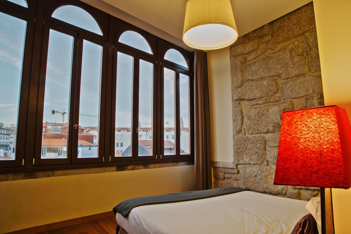 A hotel room with a bed, a large window with multiple panes, a stone wall, and a red lamp.