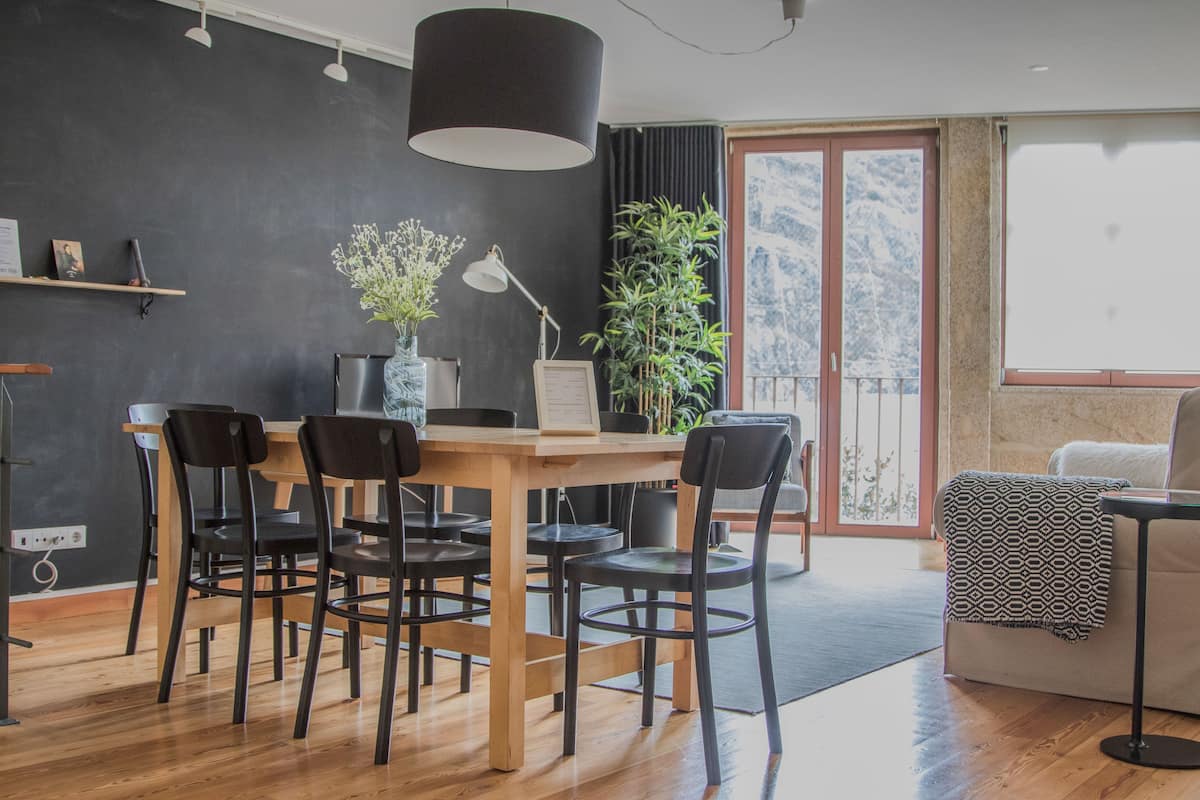 A modern living room with a wooden dining table, black chairs, a black lamp, and a large window with a view of mountains.