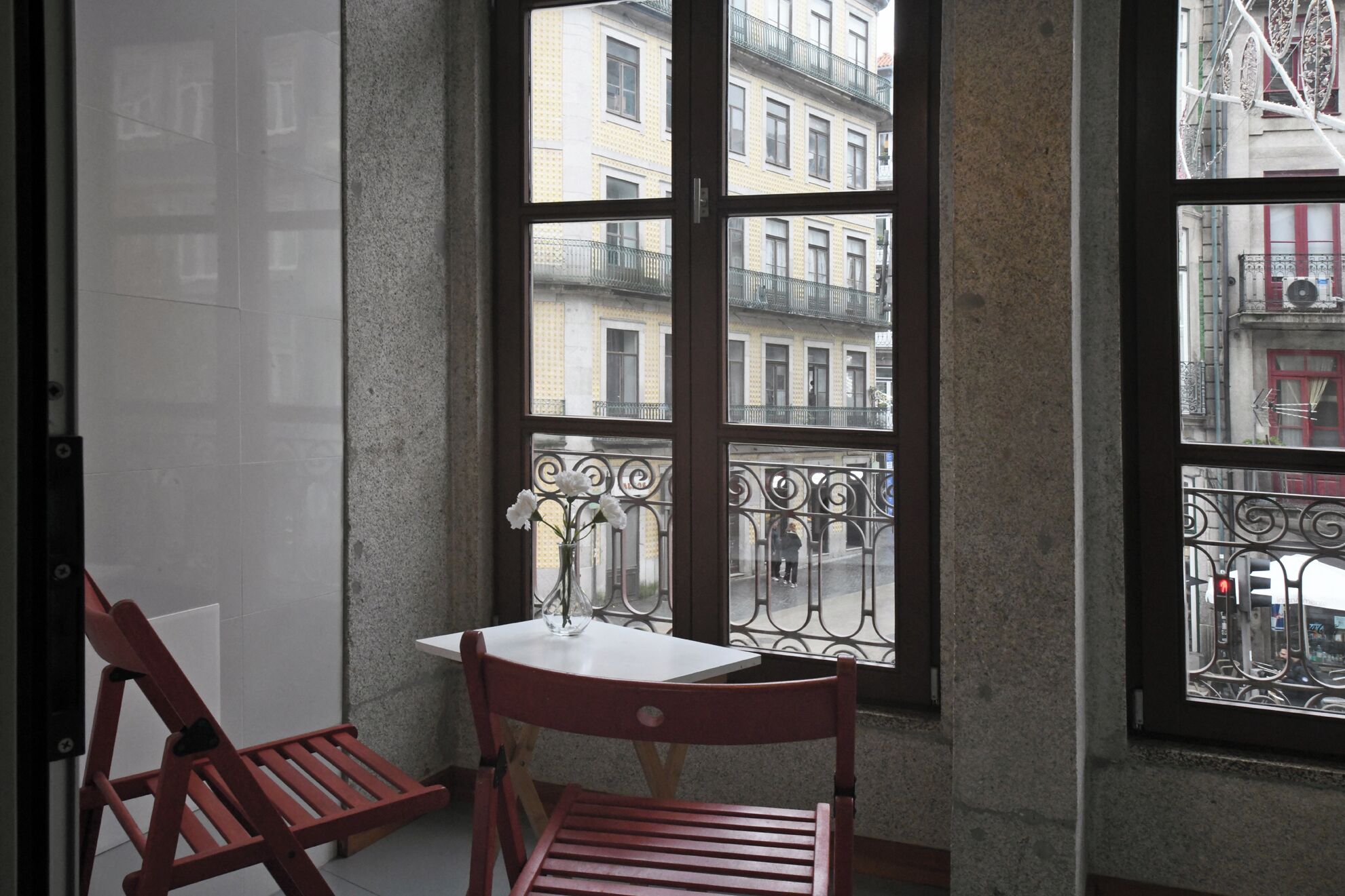 Quarto de canto com uma mesa e duas cadeiras, janela com vista para edifícios e varanda com guarda-corpo.