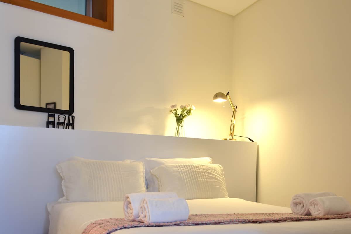A neatly made bed with white linens and a pink and white striped blanket, a bedside lamp, and a small vase with flowers on the nightstand.