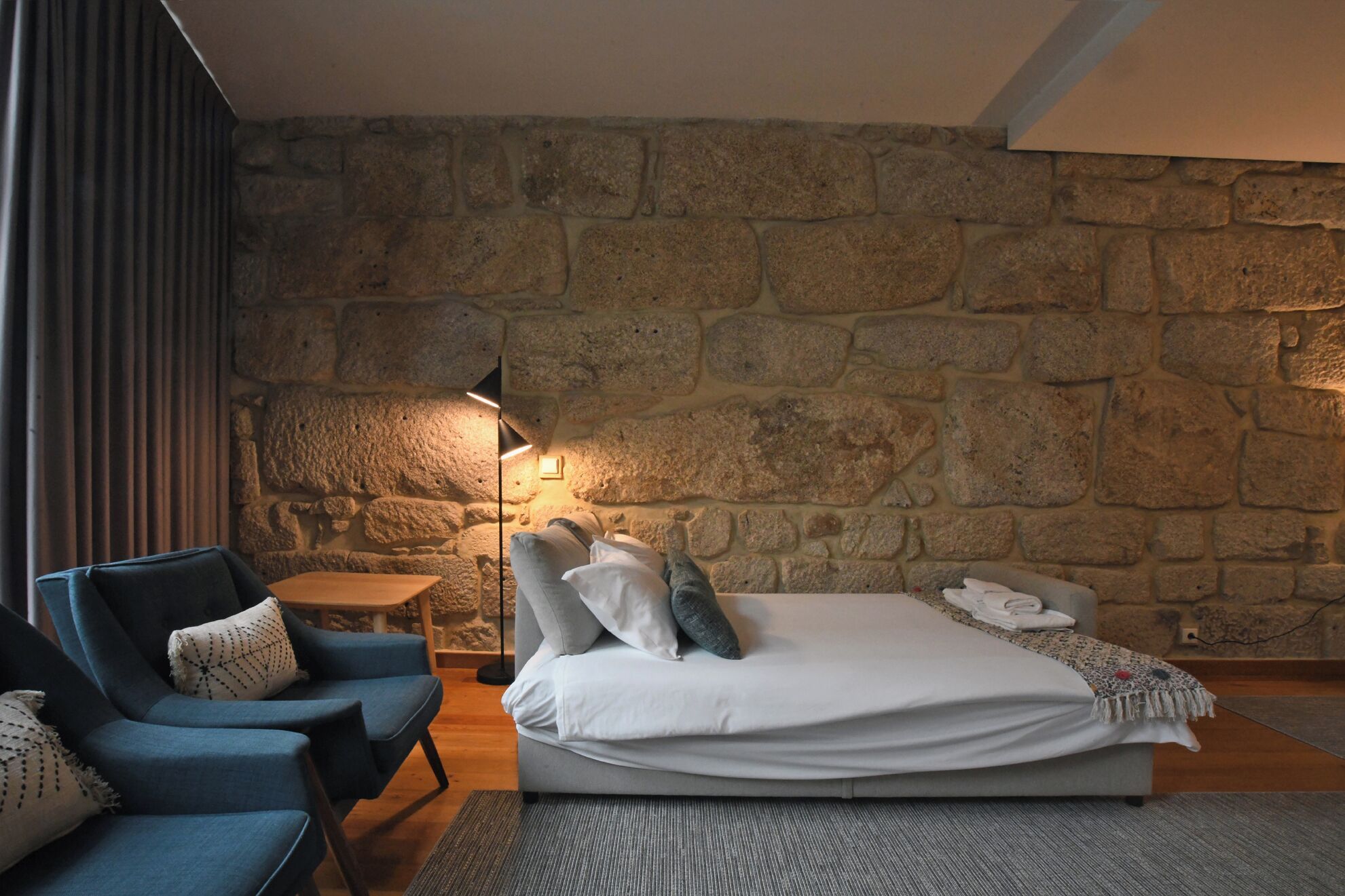 A bedroom with a stone wall, a bed with pillows, a nightstand, a lamp, and a blue chair.
