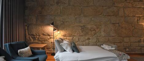 A bedroom with a stone wall, a bed with pillows, a nightstand, a lamp, and a blue chair.