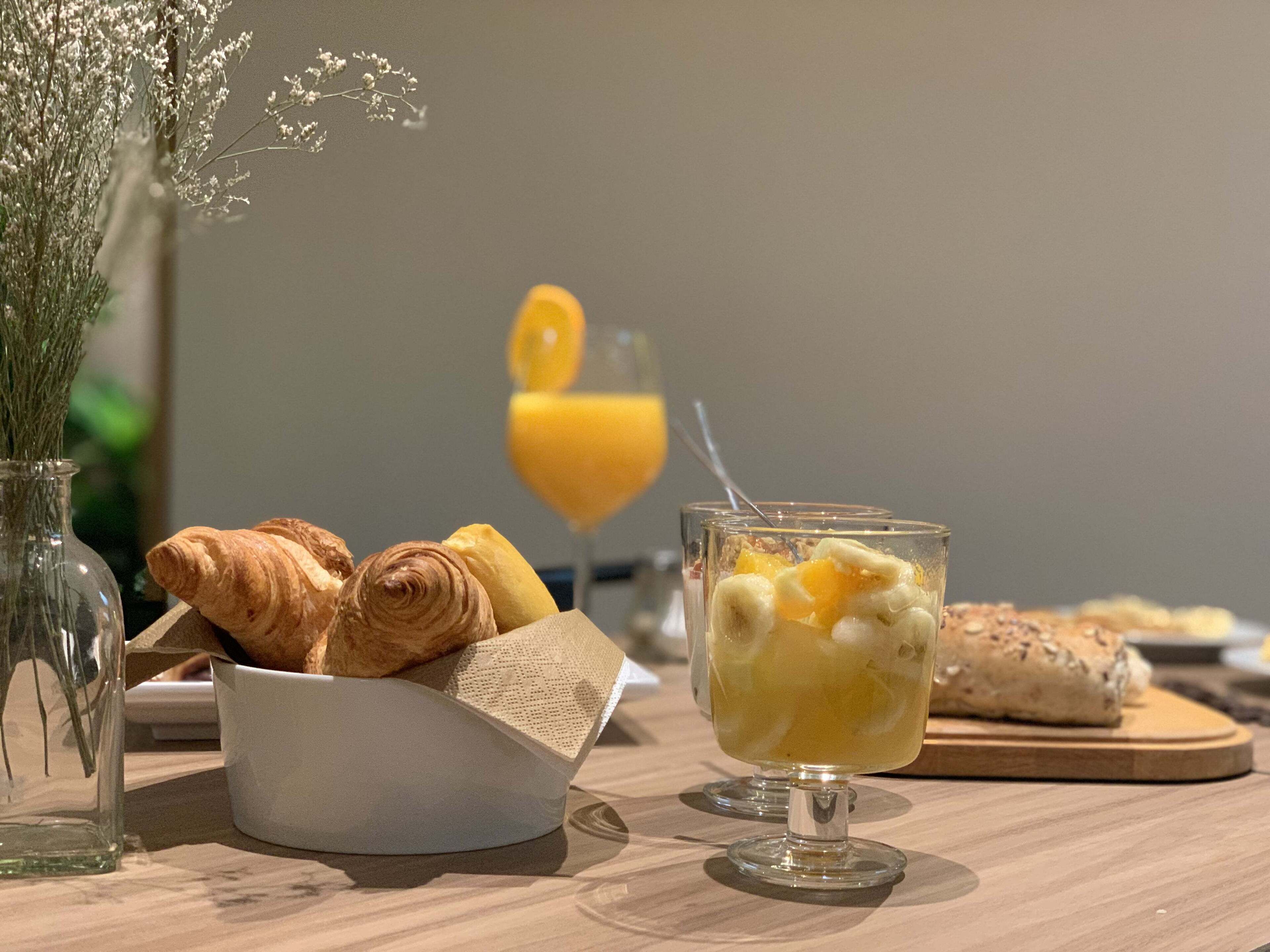 Desayuno con cruasanes, un bol de fruta y un vaso de jugo de naranja sobre una mesa de madera.