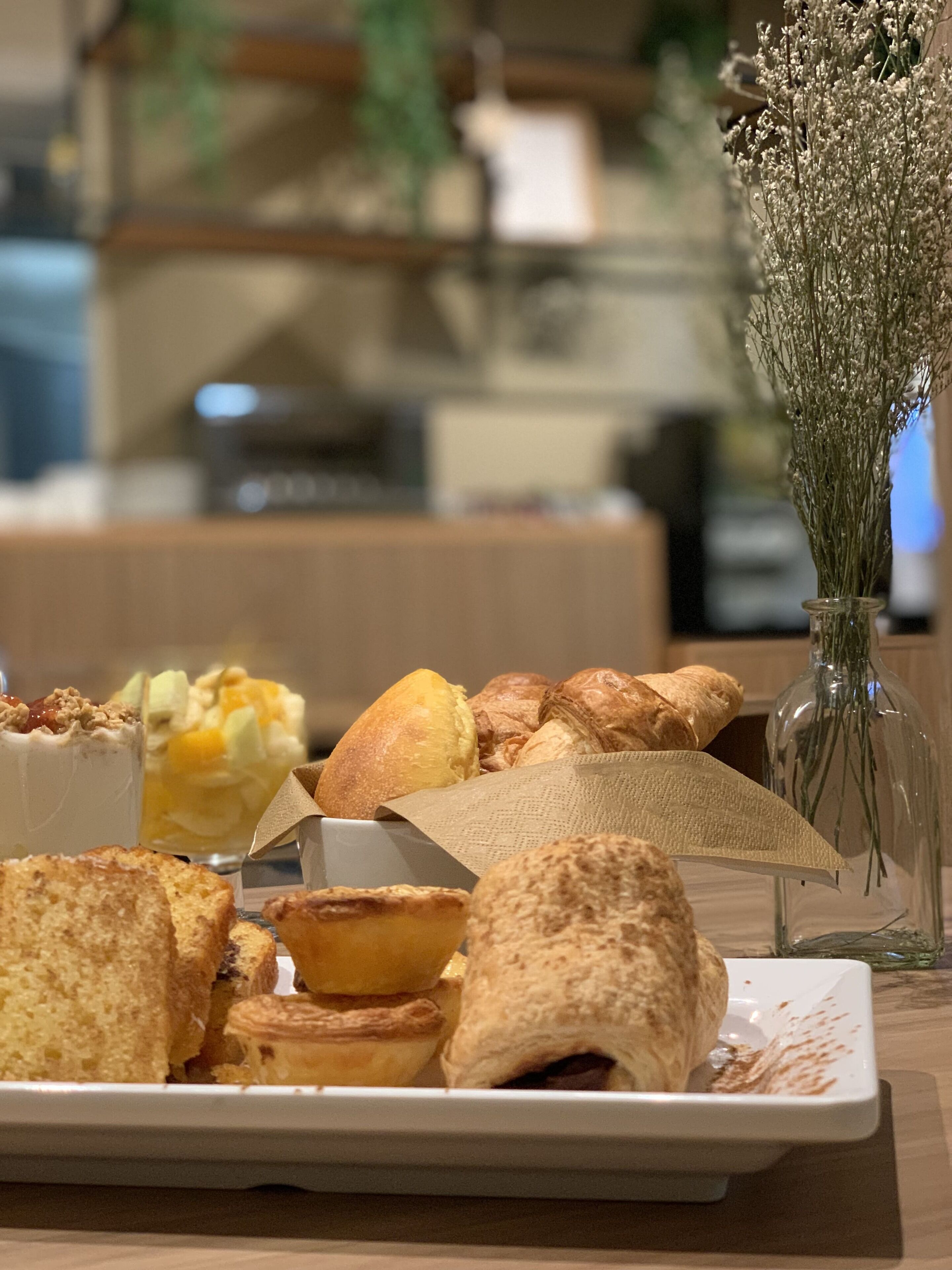 A variety of pastries and desserts on a white plate, with a vase of dried flowers in the background.