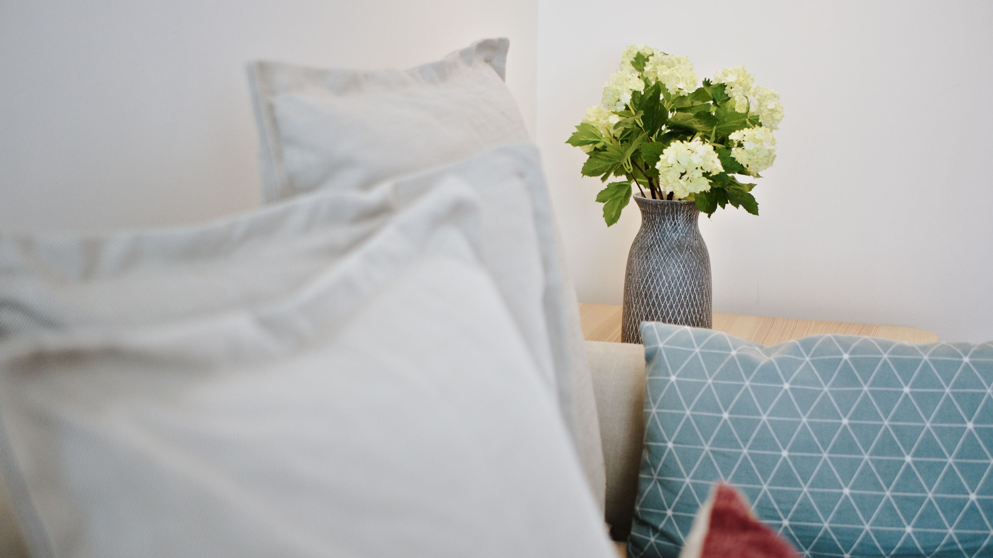 Una cama con almohadas de estampado blanco y azul, un florero con flores blancas y un florero texturizado sobre una mesita de noche de madera.