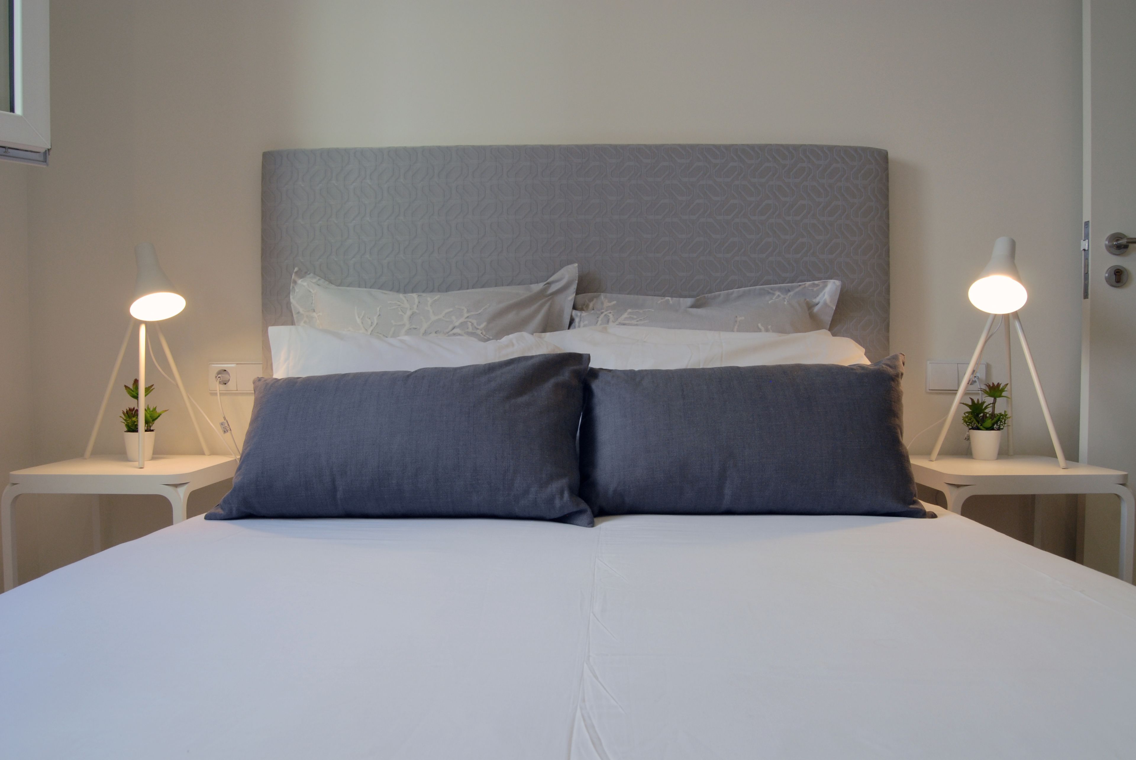 Una cama bien hecha con ropa de cama blanca y gris, dos mesitas de noche con lámparas, y una pequeña planta en maceta sobre cada mesita.
