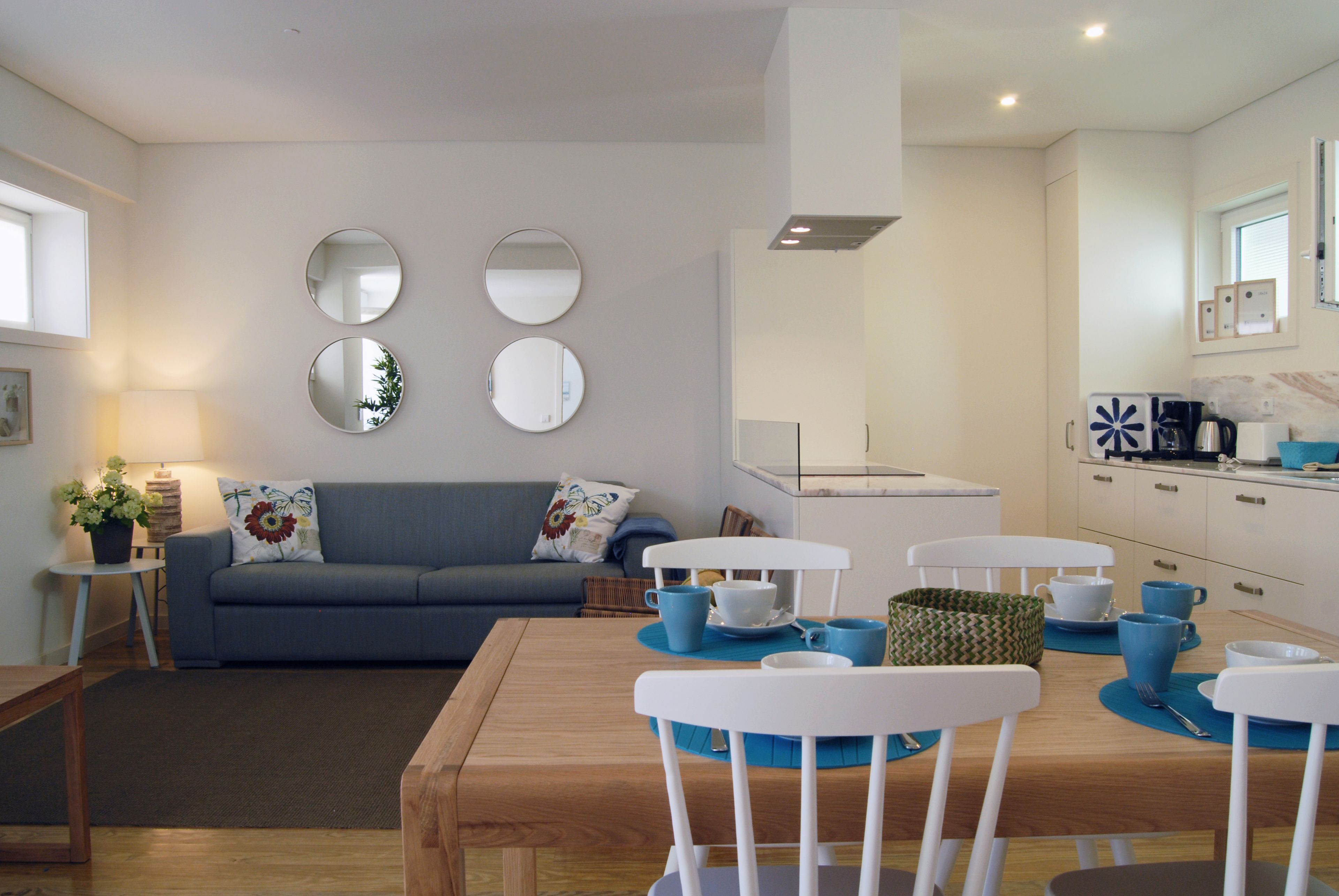 A modern living room with a blue sofa, a wooden dining table, and white chairs. There are two round mirrors on the wall, a small side table with a lamp, and a kitchen area visible in the background.
