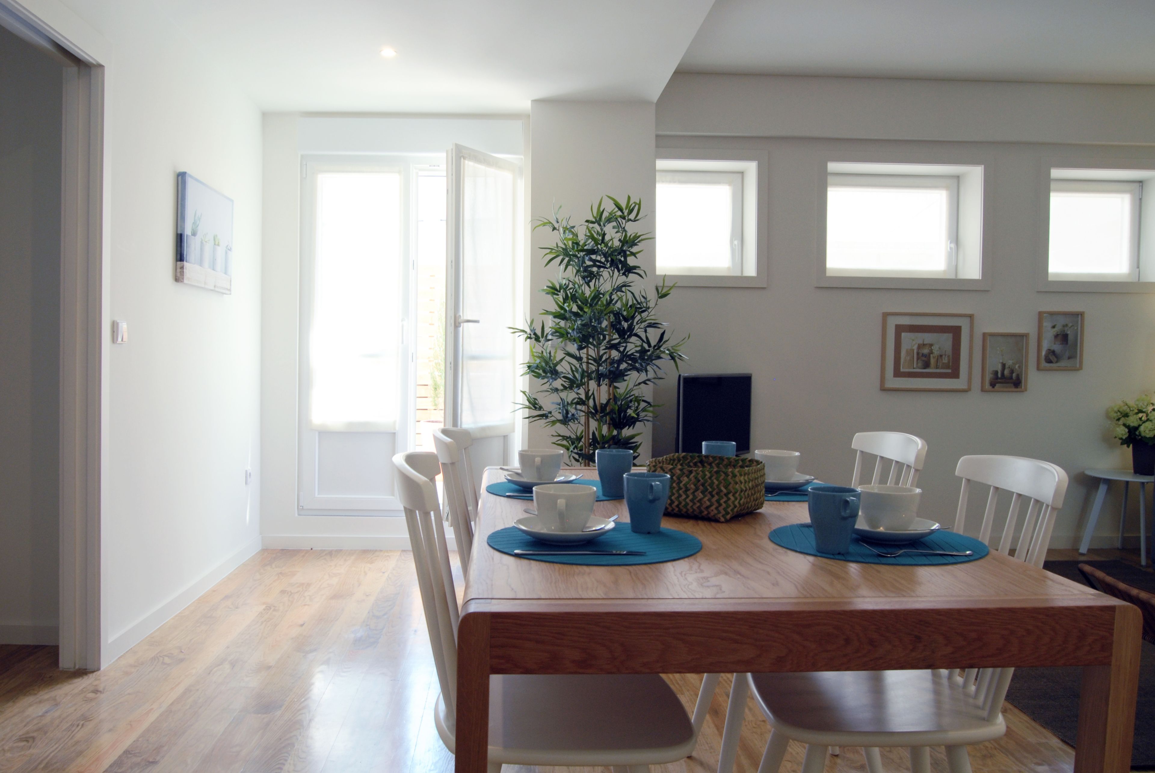A dining area with a wooden table, white chairs, blue placemats, and a potted plant.
