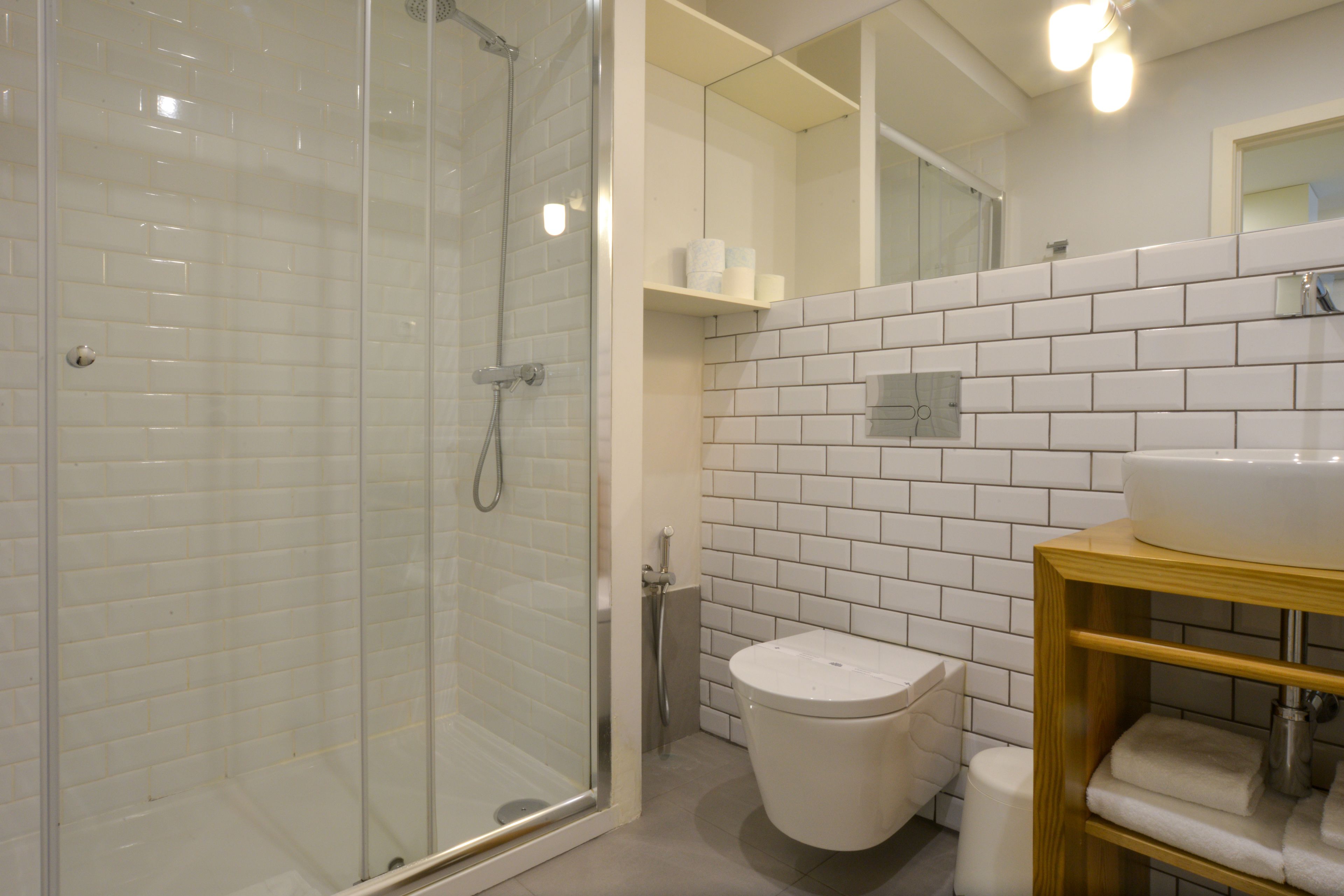 A modern bathroom with a glass-enclosed shower, white tiled walls, a wooden vanity, and a toilet.