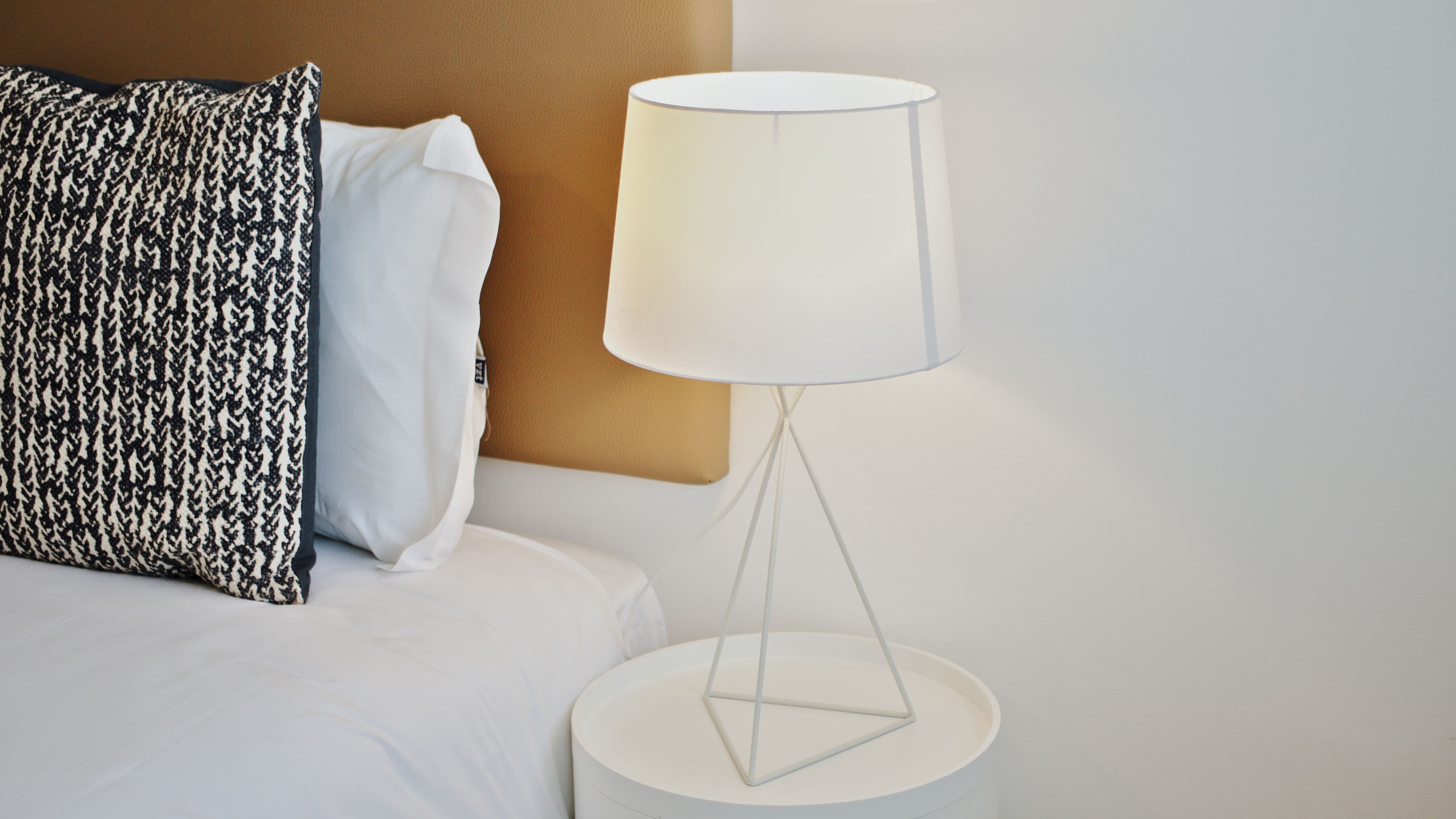 Un letto con biancheria bianca, un cuscino a motivi bianchi e neri e una lampada bianca con base geometrica su un comodino bianco.