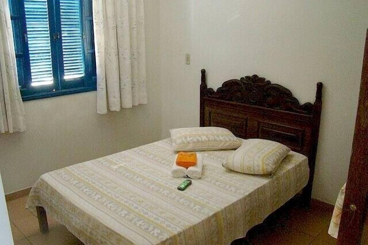 Un dormitorio con cabecera de madera, cama con ropa de cama estampada, ventana con cortinas y suelo de baldosas.
