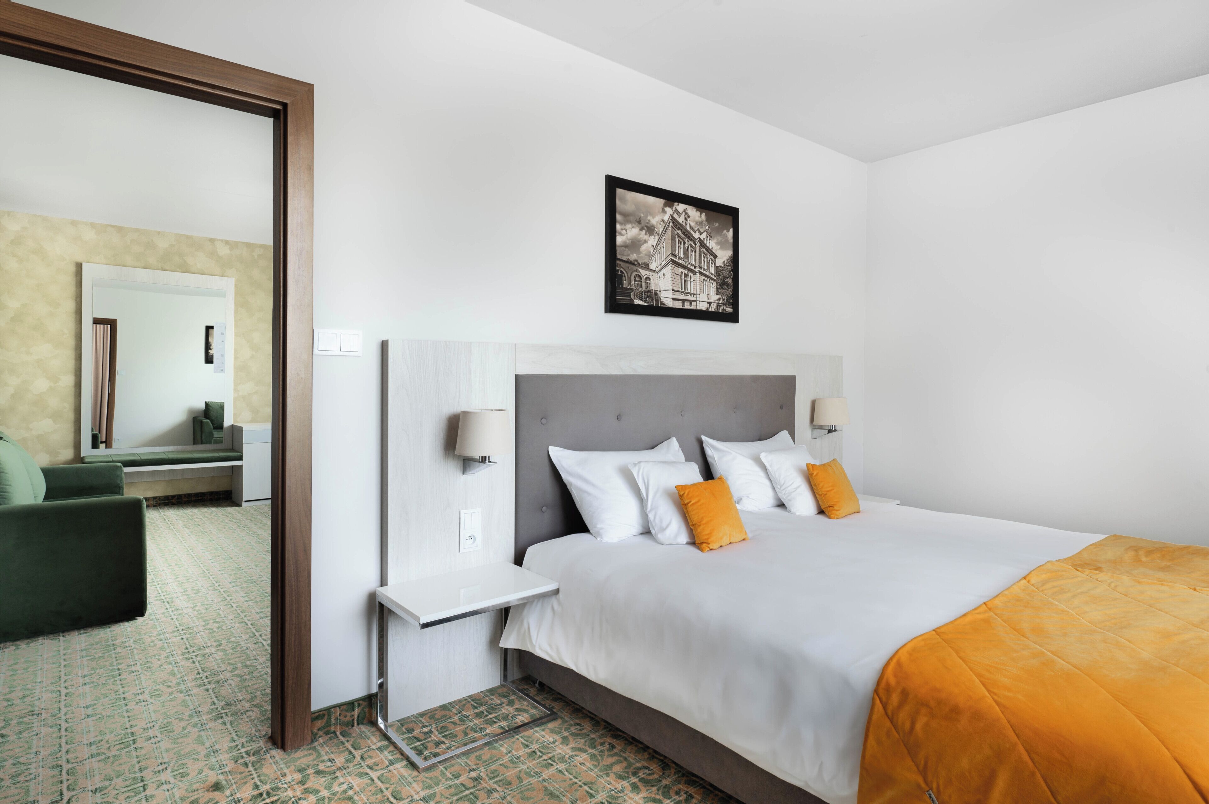 Una habitación de hotel con cama, mesitas de noche, un espejo, un cuadro en la pared y una silla verde.