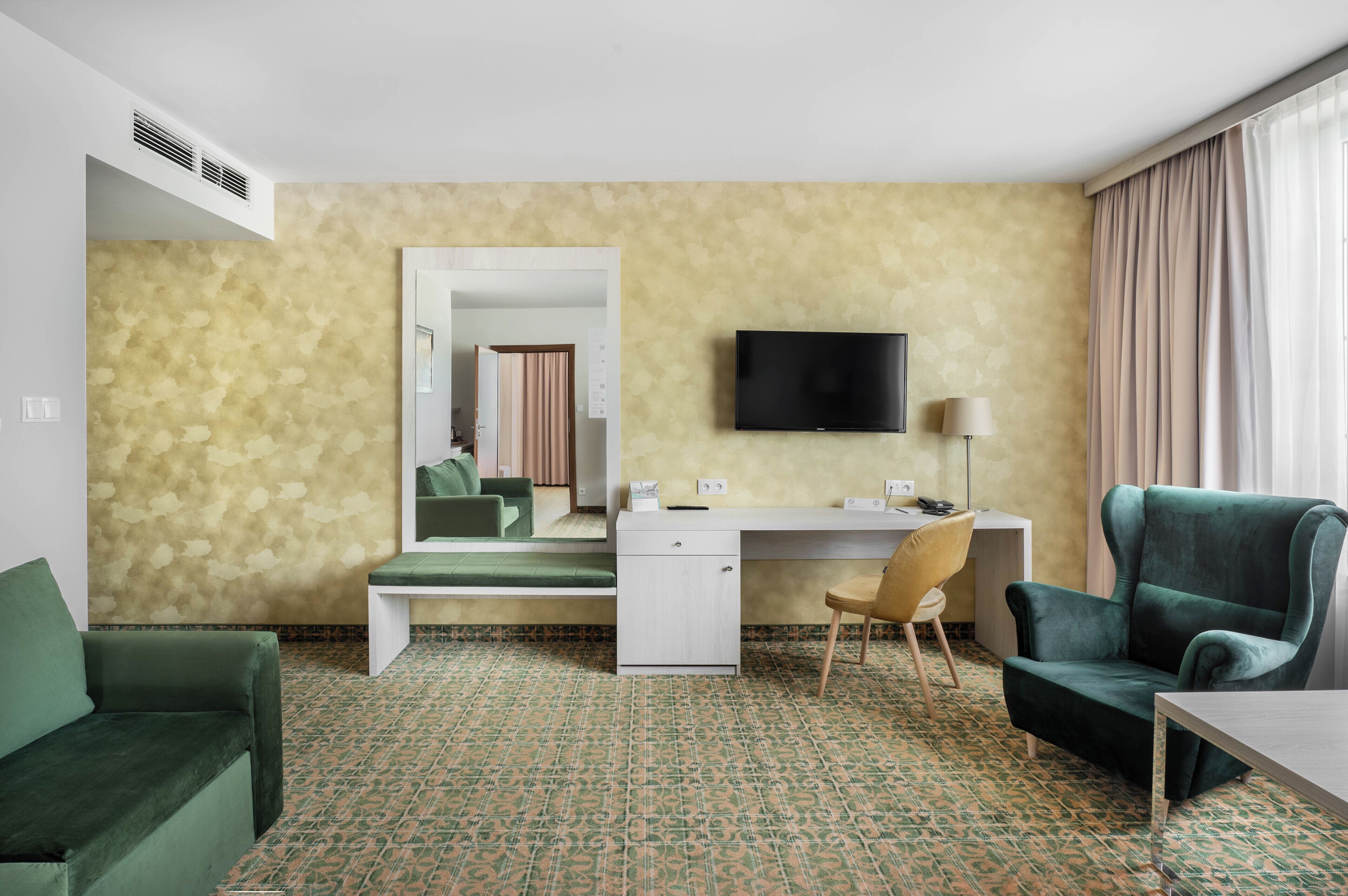 Habitación de hotel con sofá verde, un escritorio, una silla, un televisor y un espejo grande.