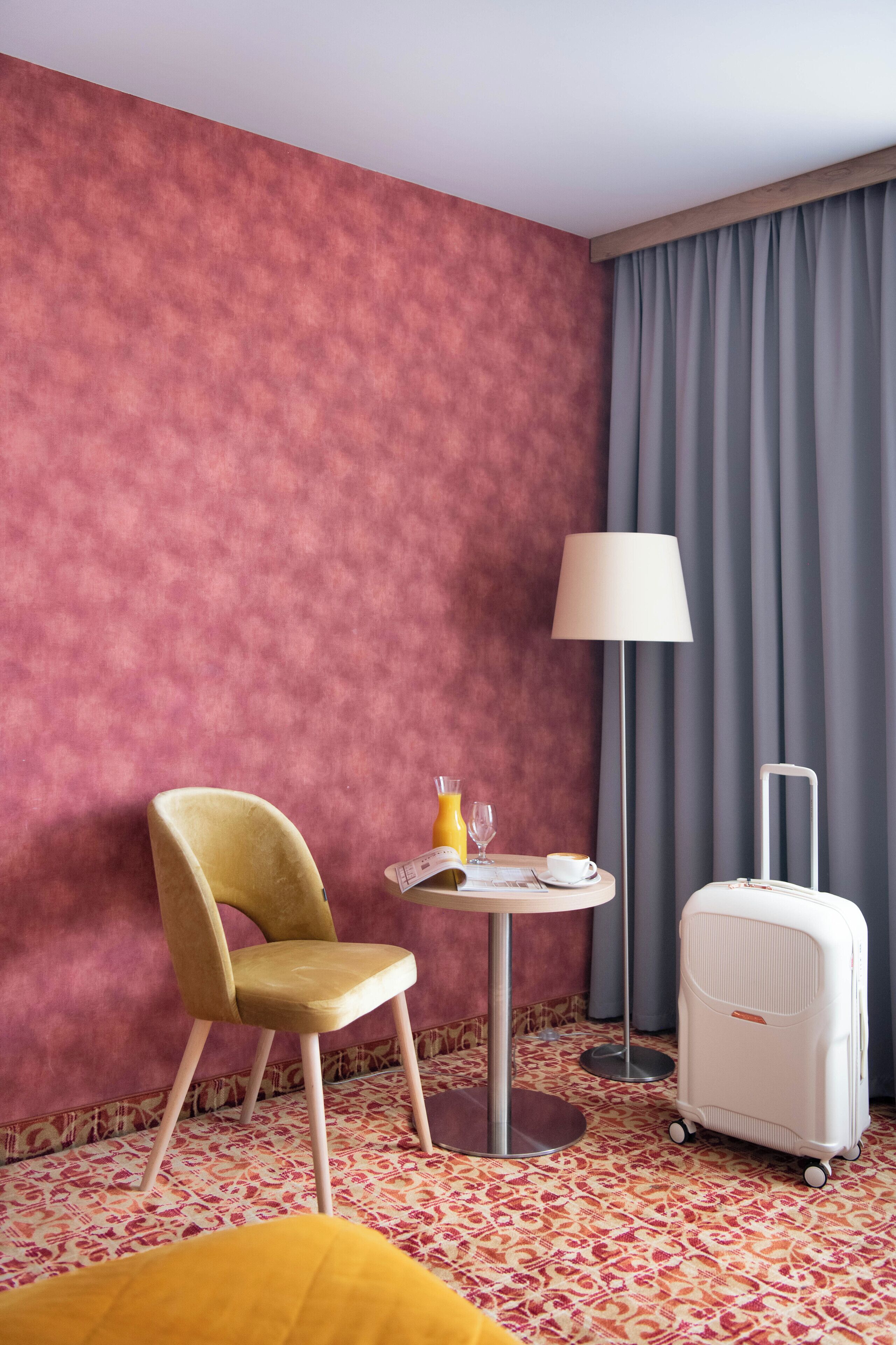 A hotel room with a patterned carpet, a yellow chair, a small round table with a lamp, a white suitcase, and a grey curtain.