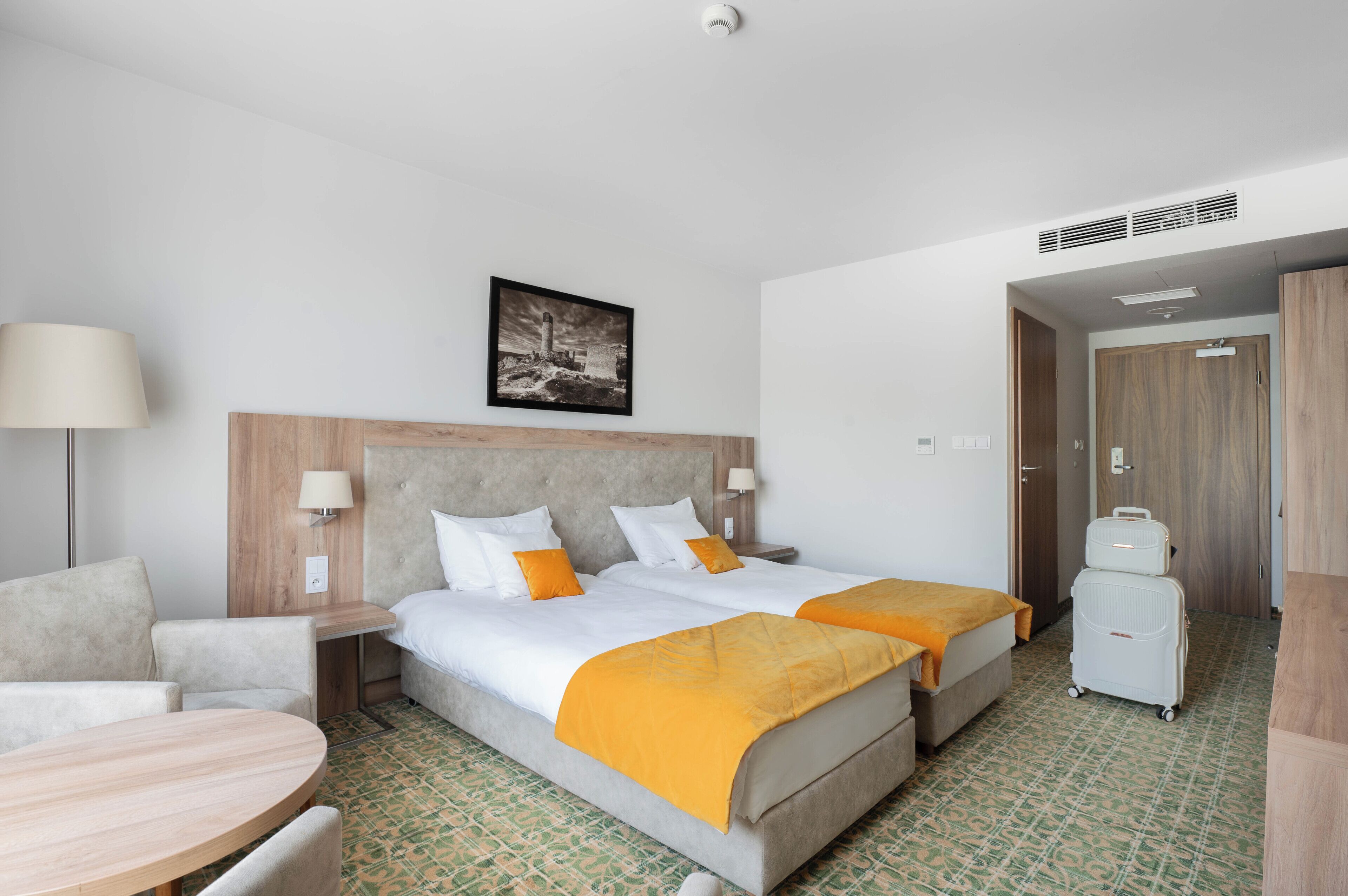 A hotel room with a double bed, two bedside tables, a desk, and a chair. There is a suitcase on the floor.