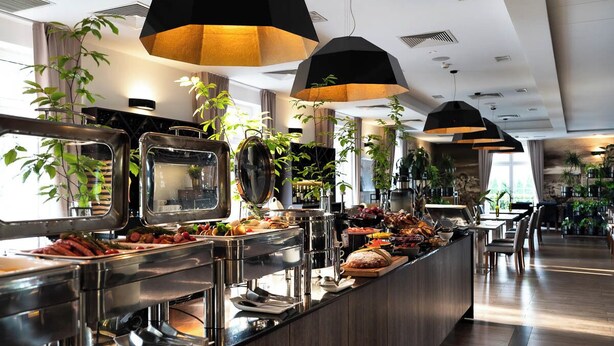 Zona de bufé con diversas opciones de comida, que incluyen frutas, verduras y carnes. Hay varias estaciones de servicio con mostradores de acero inoxidable e iluminación colgante negra.
