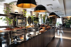 Zona de bufé con diversas opciones de comida, que incluyen frutas, verduras y carnes. Hay varias estaciones de servicio con mostradores de acero inoxidable e iluminación colgante negra.
