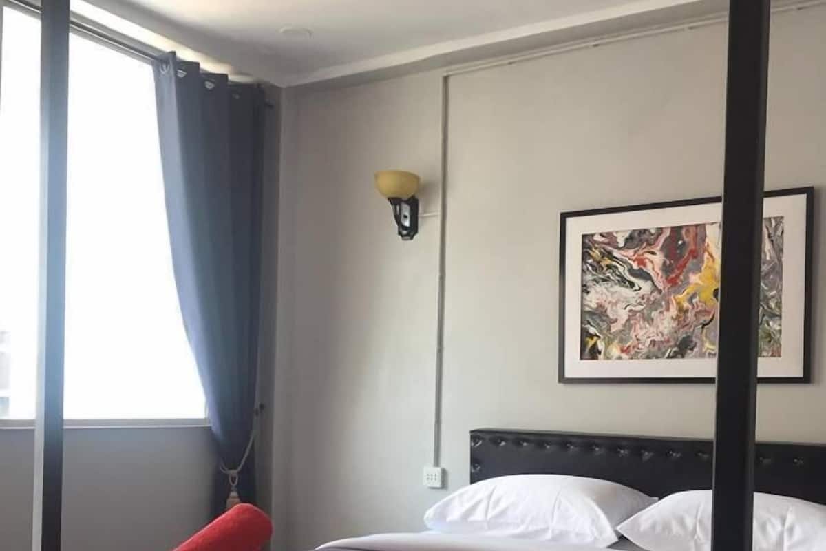 A neatly made bed with white and gray bedding, a red towel, and a black headboard. There is a framed abstract painting on the wall and a window with blue curtains.
