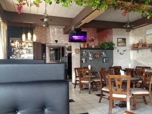 Un área de bar con sillas de madera y un banco acolchado negro. Hay plantas colgantes y una pantalla de televisión mostrando 'estbent'.
