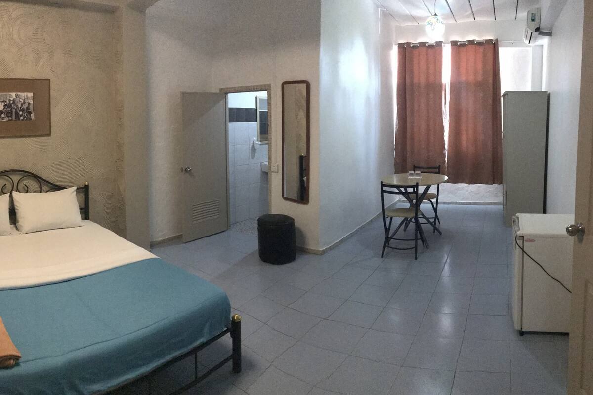 A hotel room with a bed, a small desk, a chair, a mirror, and a refrigerator.