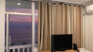 A room with a television on a wooden table, a window with a view of the sea, and curtains. - Seaview (Pattaya)