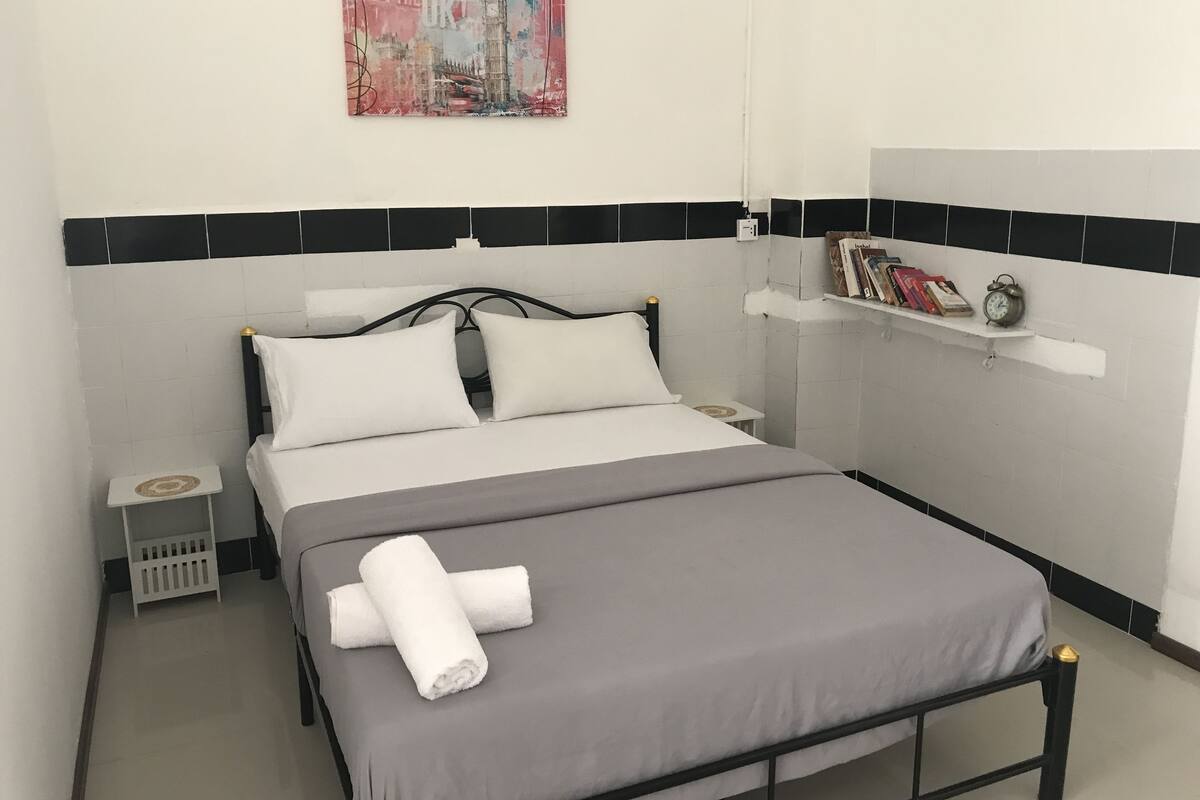 A neatly made bed with a grey bedspread and white pillows, a small bedside table with a lamp, and a shelf with books and a clock.