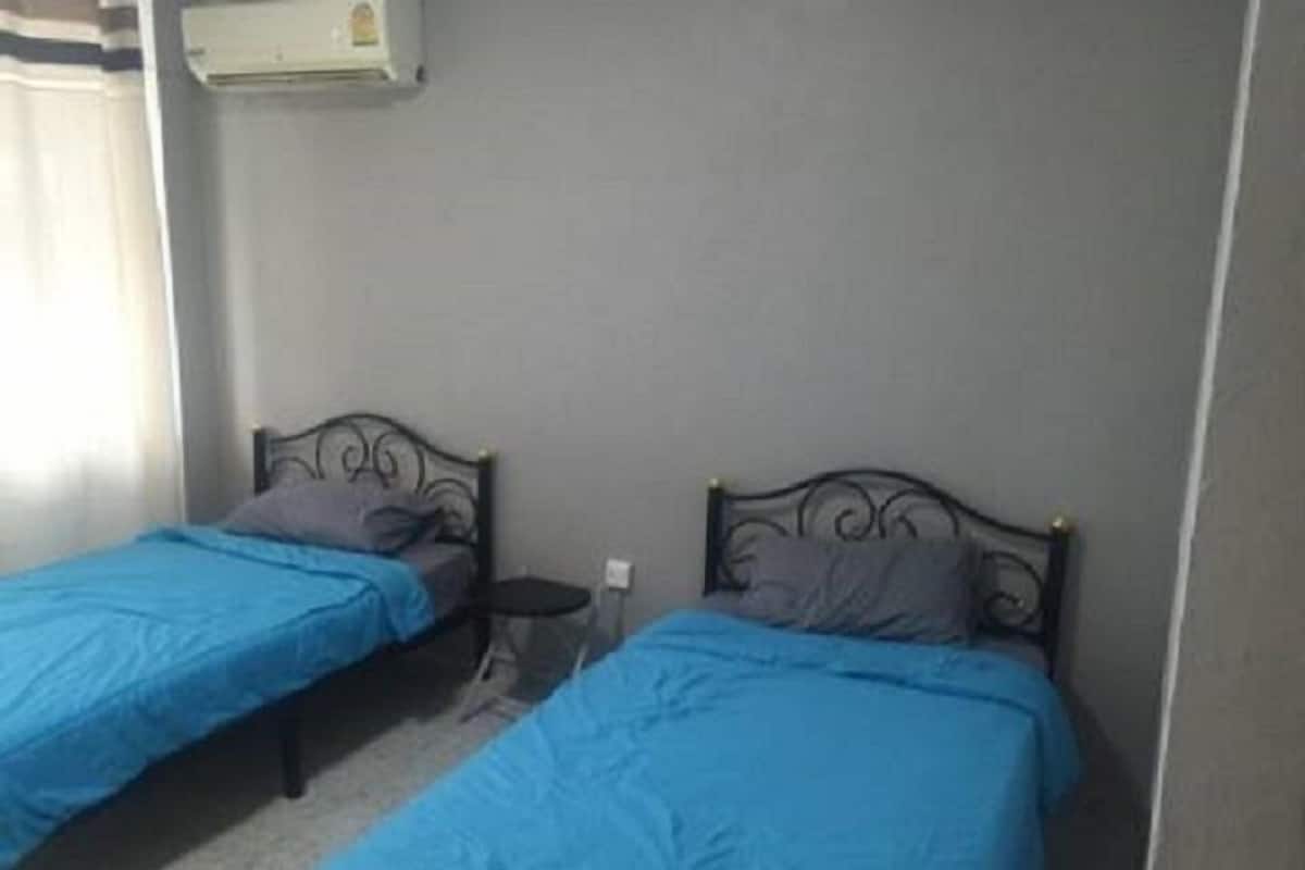 Two single beds with metal frames and blue bedding in a room with a plain wall and an air conditioning unit above.