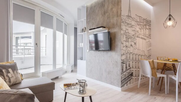 Ein modernes Wohnzimmer mit großem Fenster, einem Wand-Fernseher an einer strukturierten Wand, einem Essbereich mit Tisch und Stühlen sowie einem kleinen Beistelltisch mit Zeitschrift und Glas.